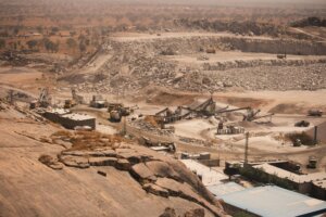 Open-pit quarry with heavy machinery and conveyor belts processing rocks in a dry, rocky landscape.