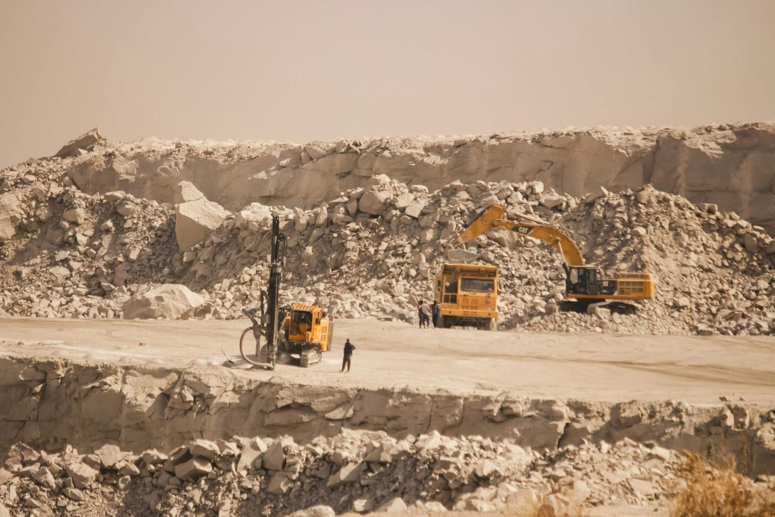 Yellow CAT excavator loading rocks into a dump truck at a quarry with workers nearby