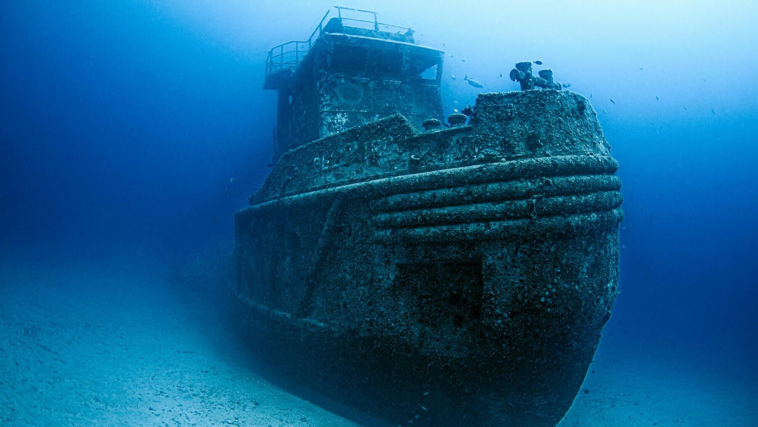 Sunken shipwreck covered in marine growth resting on the ocean floor with fish swimming nearby