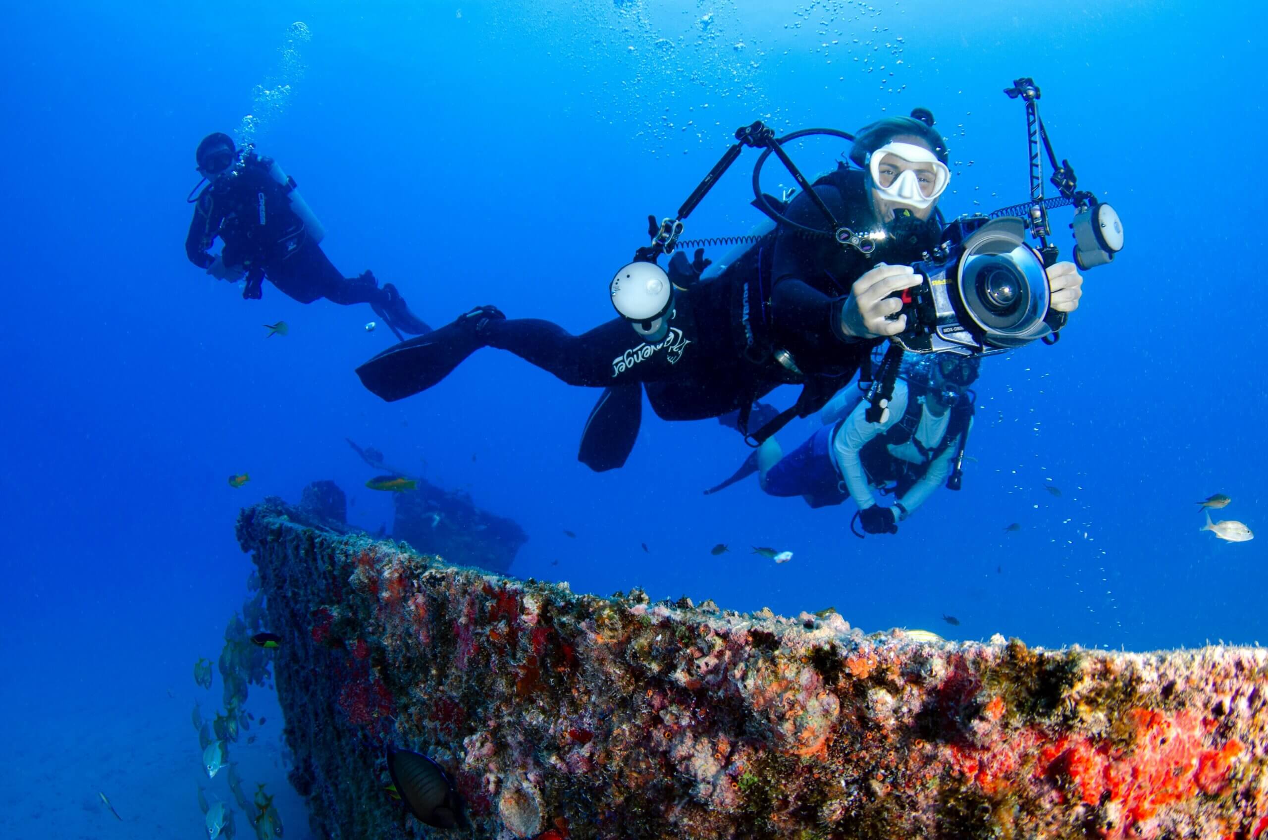Scuba divers exploring a coral-covered shipwreck, one holding an underwater camera rig.