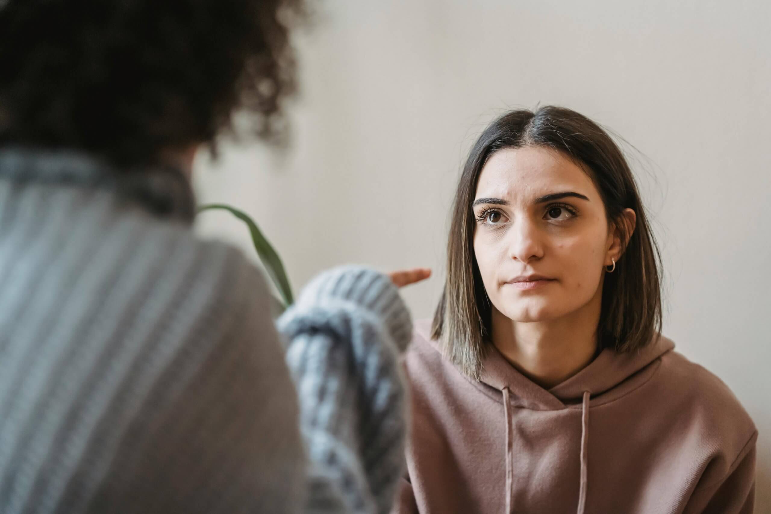 Young woman in a brown hoodie listening intently to another person pointing at her during a conversation