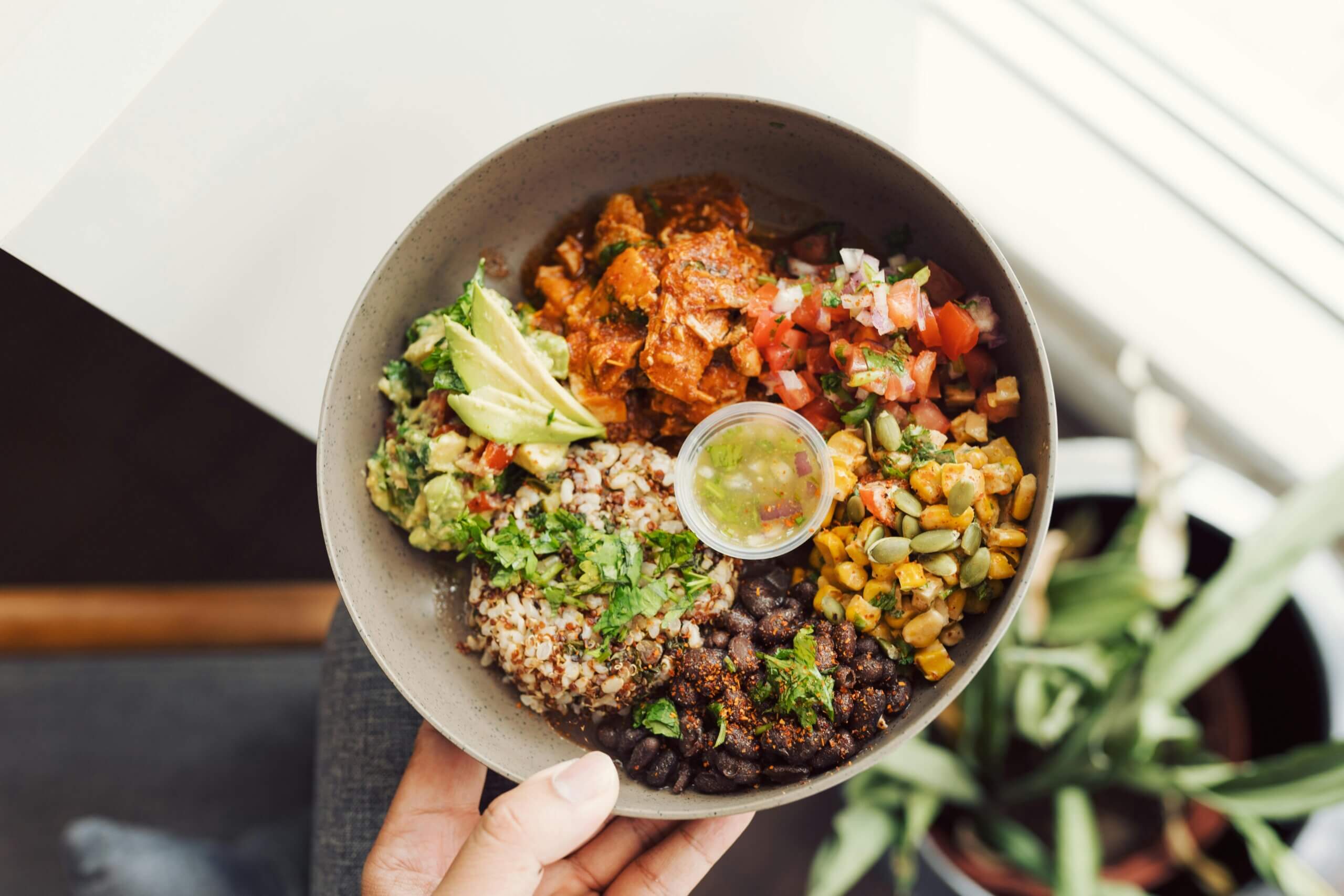 Hand holding a bowl with rice, black beans, corn, avocado, tomato salsa, and seasoned chicken with sauce.
