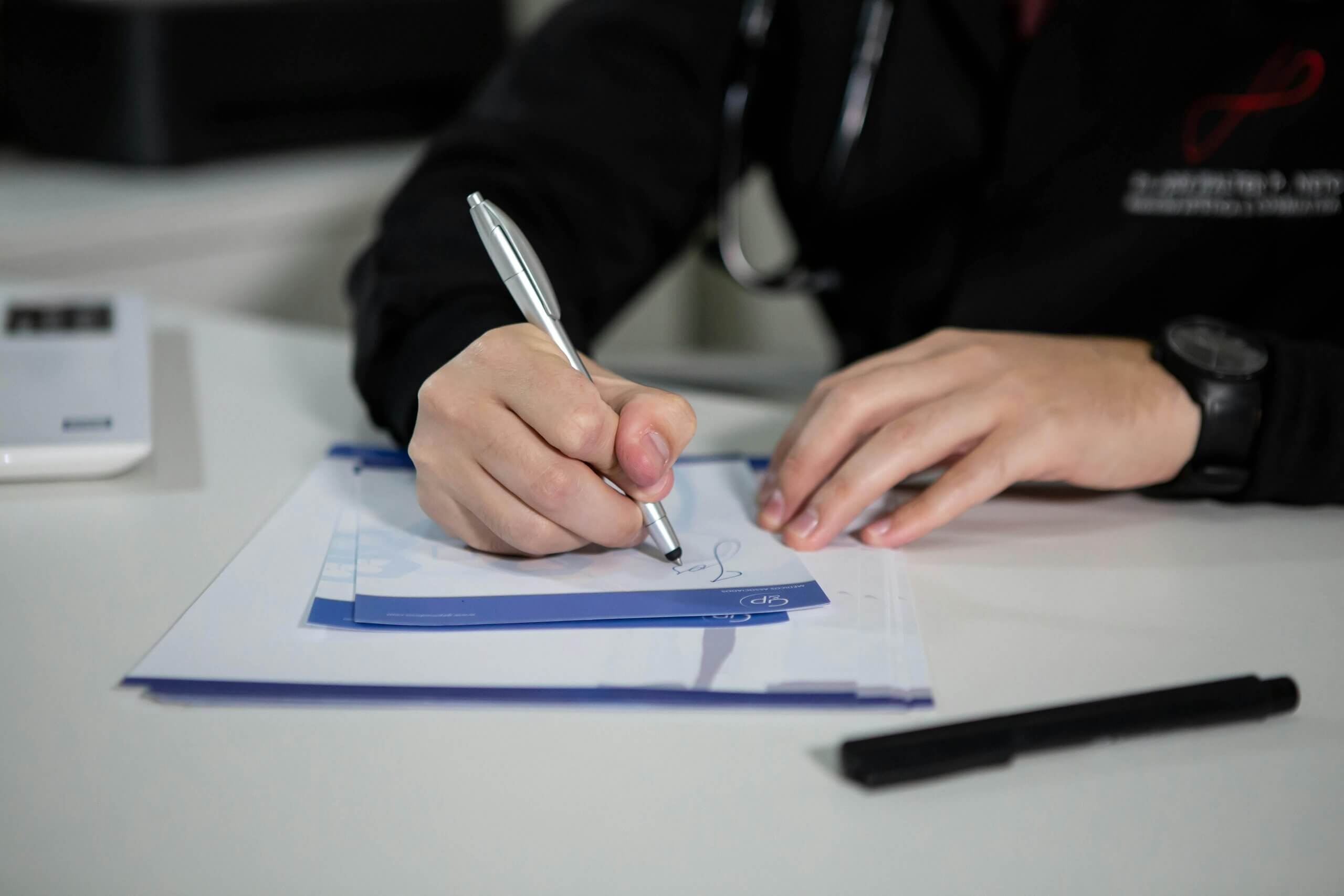 Person in black clothing signing a document with a silver pen on a white desk.