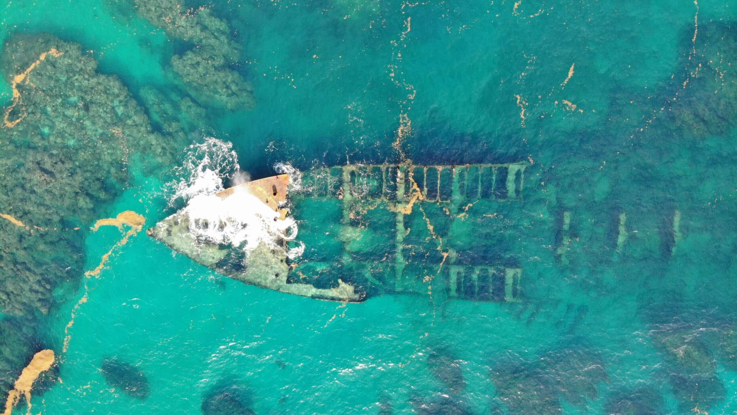 Aerial view of a partially submerged shipwreck in clear turquoise ocean water.
