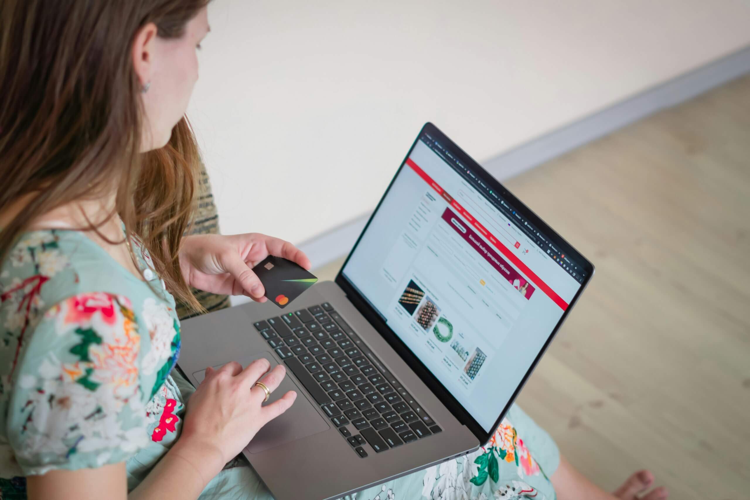 Woman holding a Mastercard credit card while shopping online on a laptop with a floral dress.