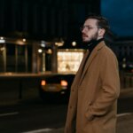 Man wearing glasses and a brown coat standing on a city street at night with blurred lights in the background.