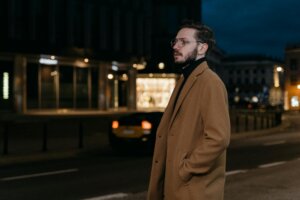 Man wearing glasses and a brown coat standing on a city street at night with blurred lights in the background.