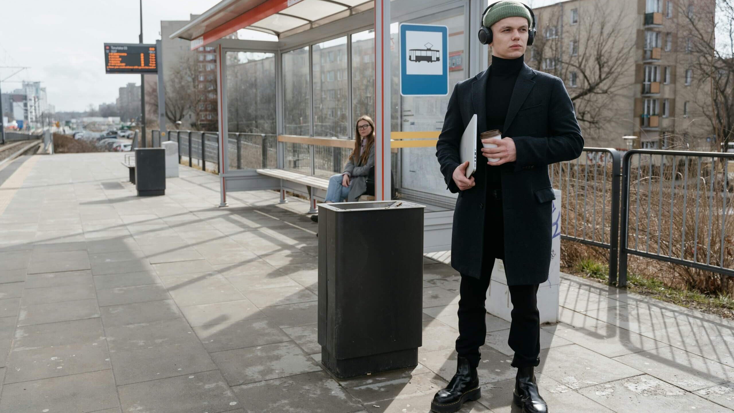 Young man in black coat and green beanie holding laptop and coffee at a tram stop with a woman sitting inside the shelter