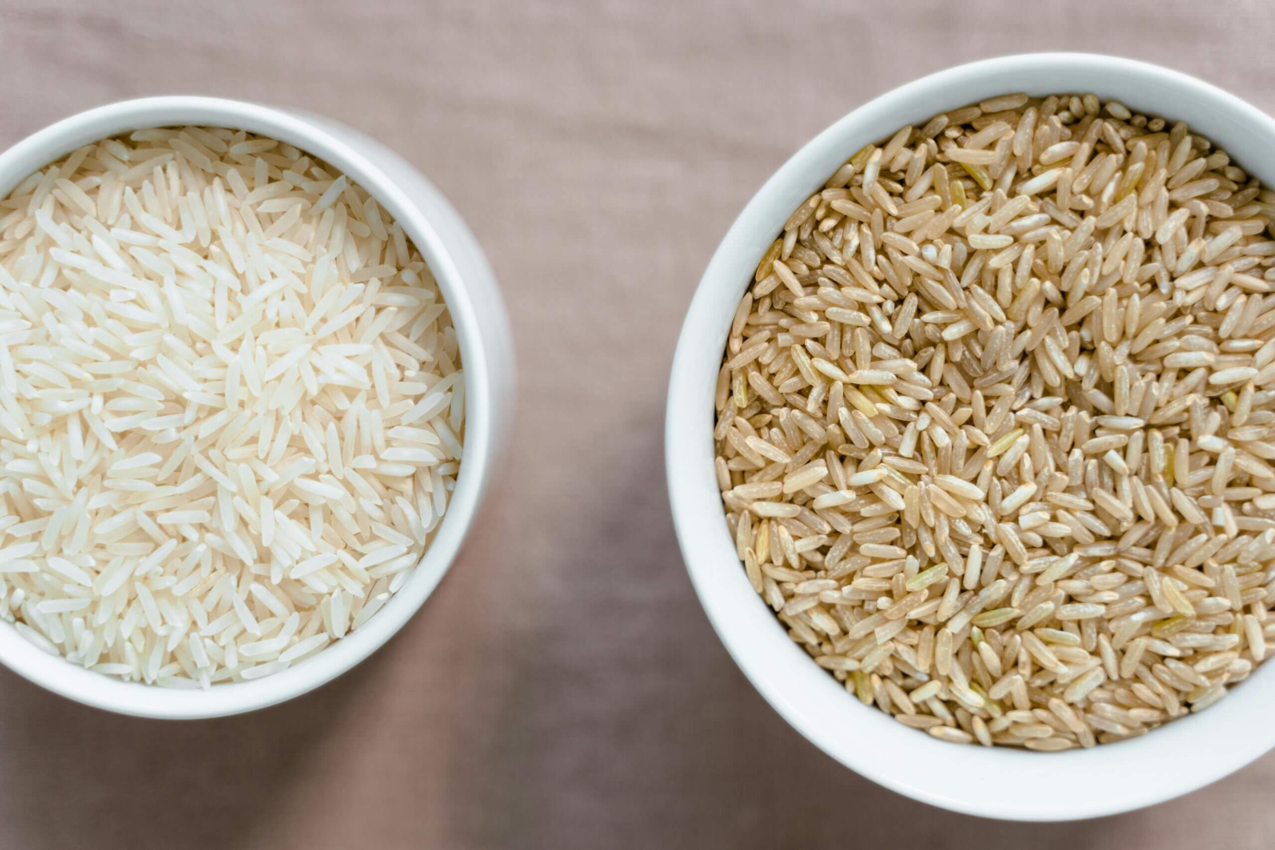 Two white bowls filled with uncooked white rice and brown rice on a beige surface