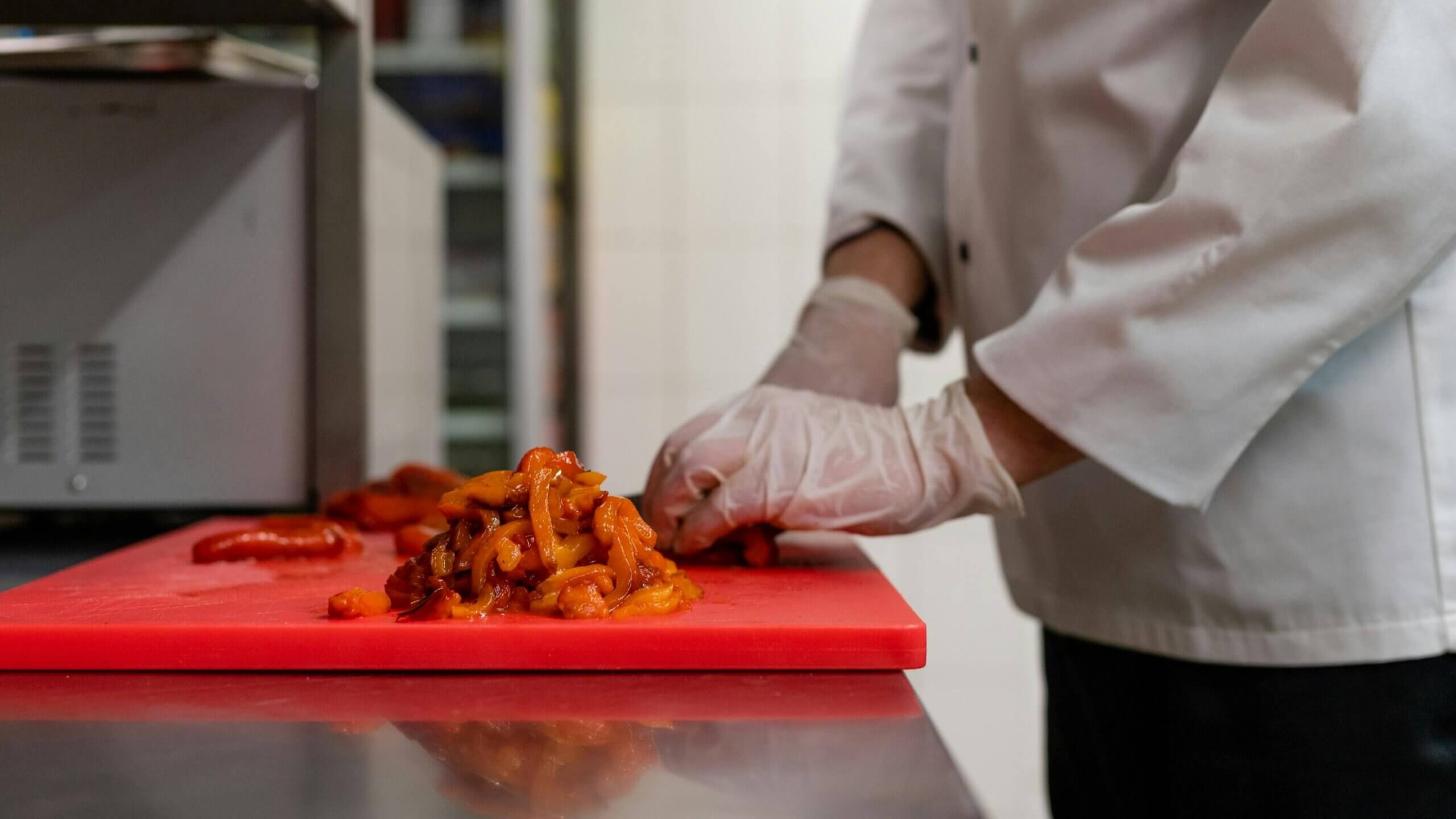 Chef wearing gloves chopping roasted red and yellow bell peppers on a red cutting board in a kitchen