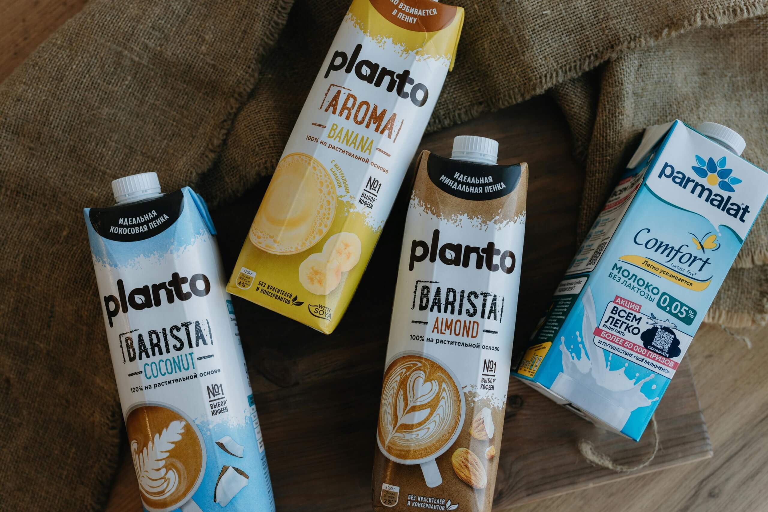 Planto Barista Coconut, Planto Aroma Banana, Planto Barista Almond, and Parmalat Comfort lactose-free milk cartons on a wooden surface.