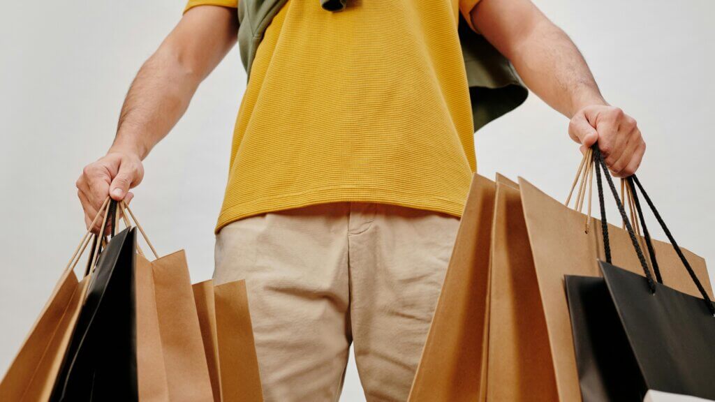 Person wearing yellow shirt and beige pants holding multiple brown and black shopping bags