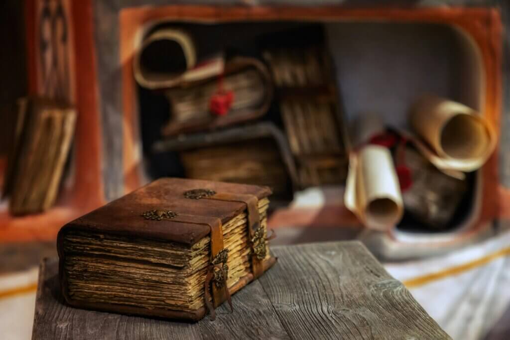 Old leather-bound book with metal clasps on a wooden table, with rolled scrolls and books in the background