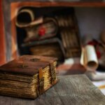 Old leather-bound book with metal clasps on a wooden table, with rolled scrolls and books in the background