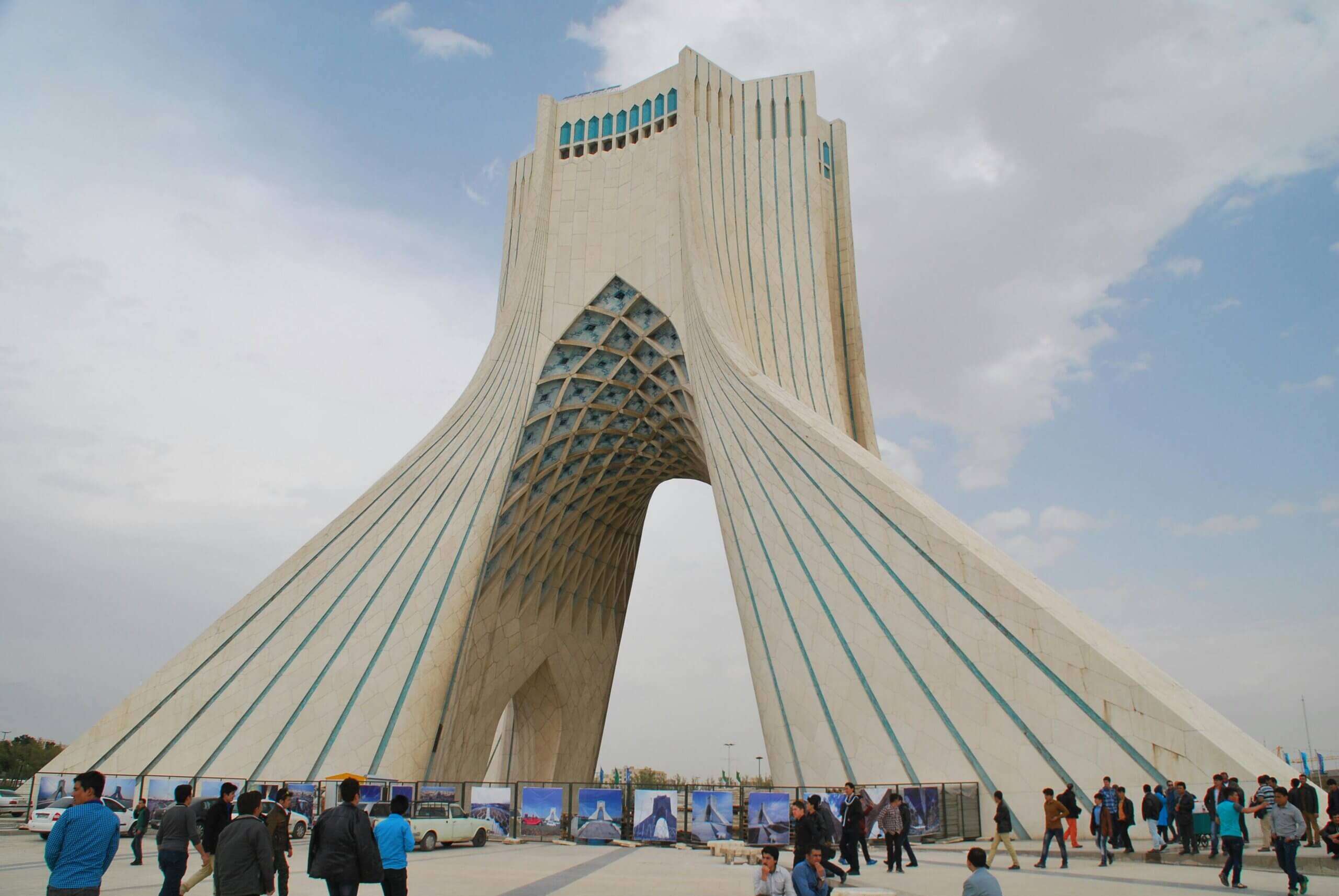 Azadi Tower in Tehran with people walking and cars parked around the base on a partly cloudy day