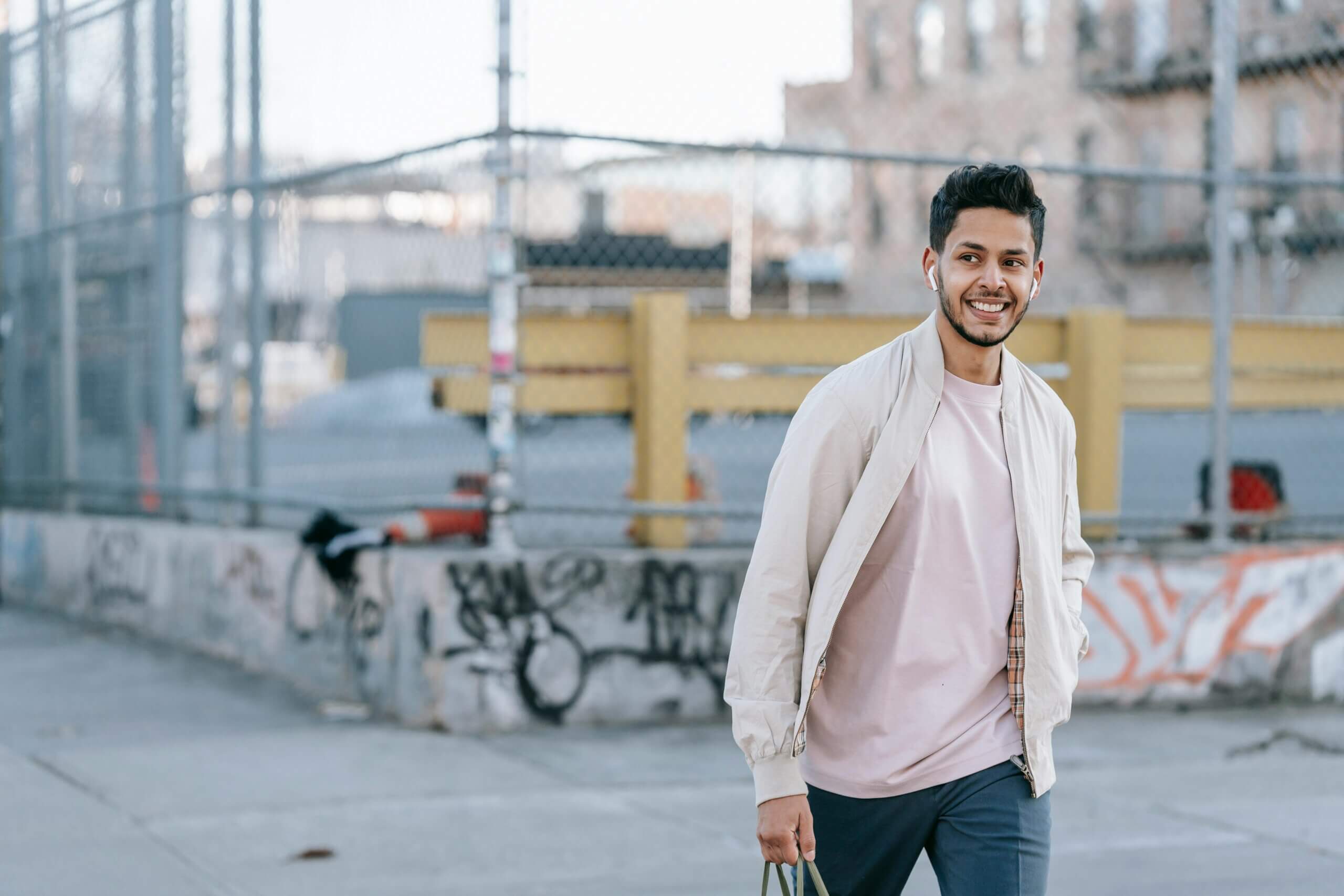 Smiling man wearing beige jacket and pink shirt walking outdoors near a graffiti-covered fence.
