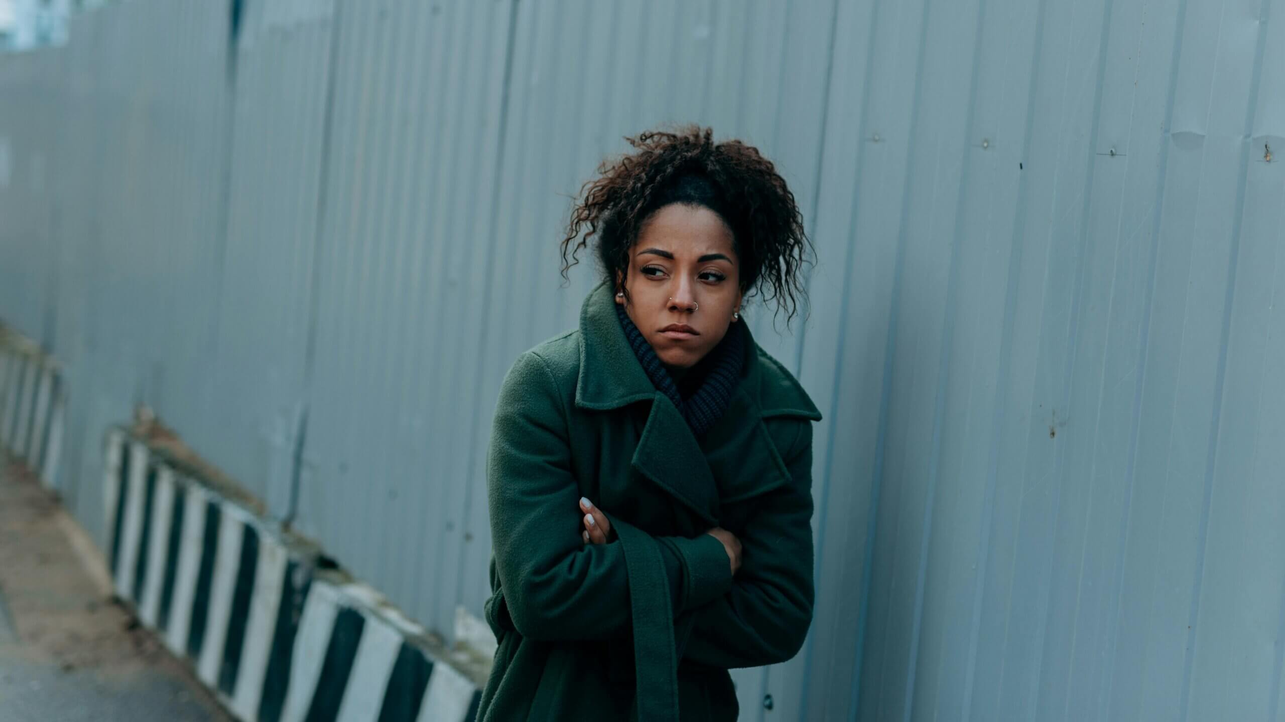 Woman in green coat standing outside with arms crossed, looking away with a serious expression.