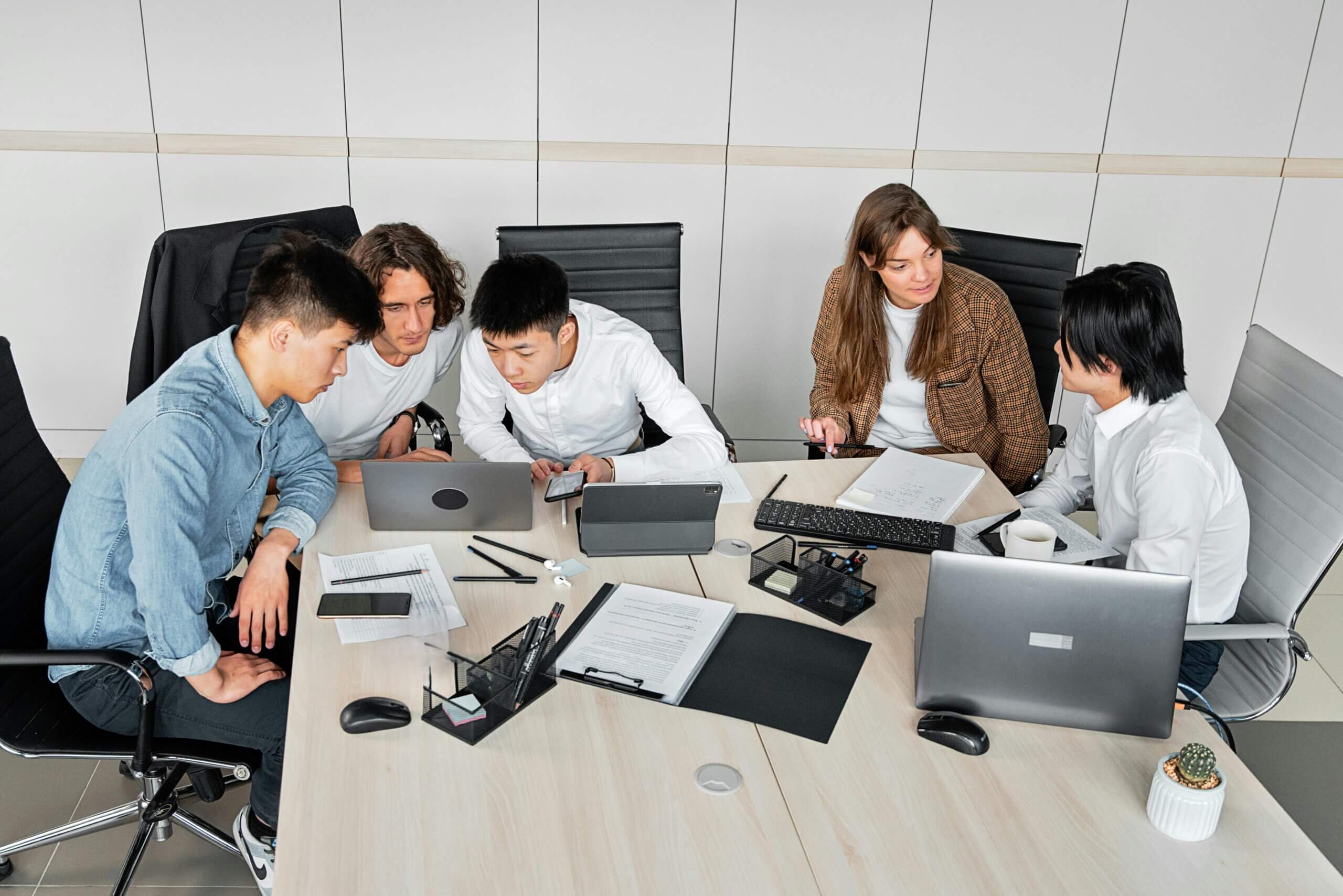 Five young professionals collaborating around a table with laptops, tablets, and documents in an office.