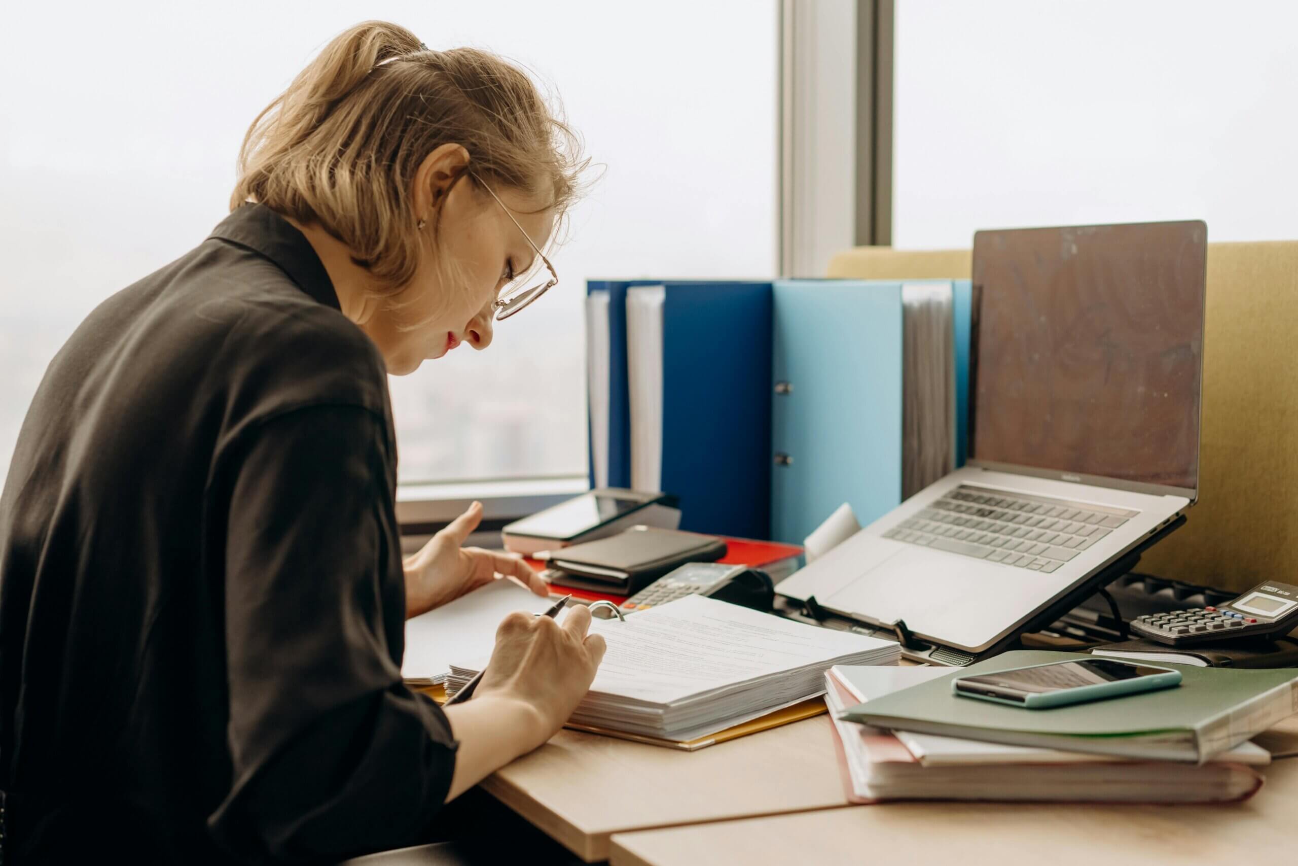 Woman in glasses and black shirt writing in a binder at a cluttered office desk with laptop and folders