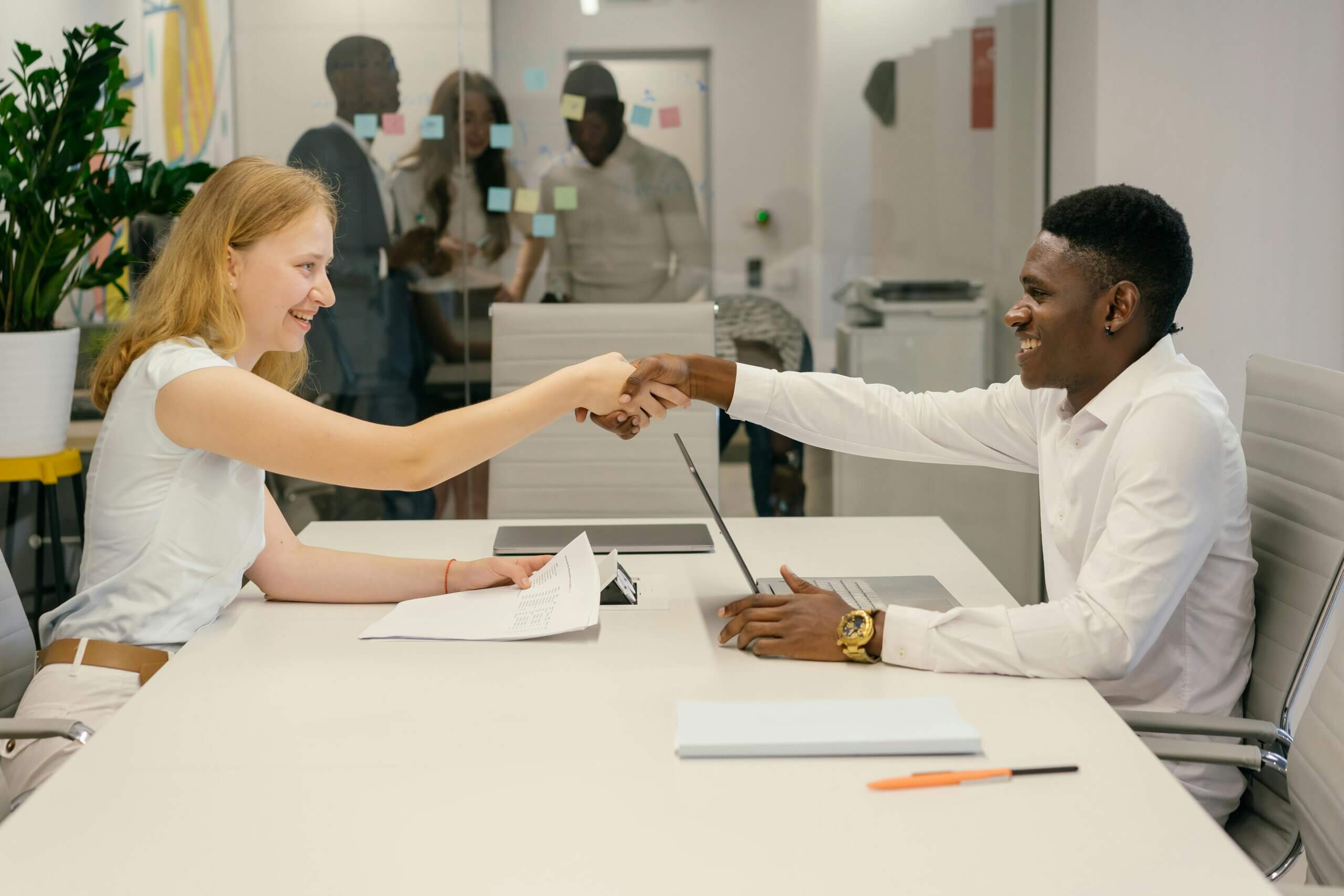 Two colleagues shaking hands across a desk in a modern office with a laptop and documents.