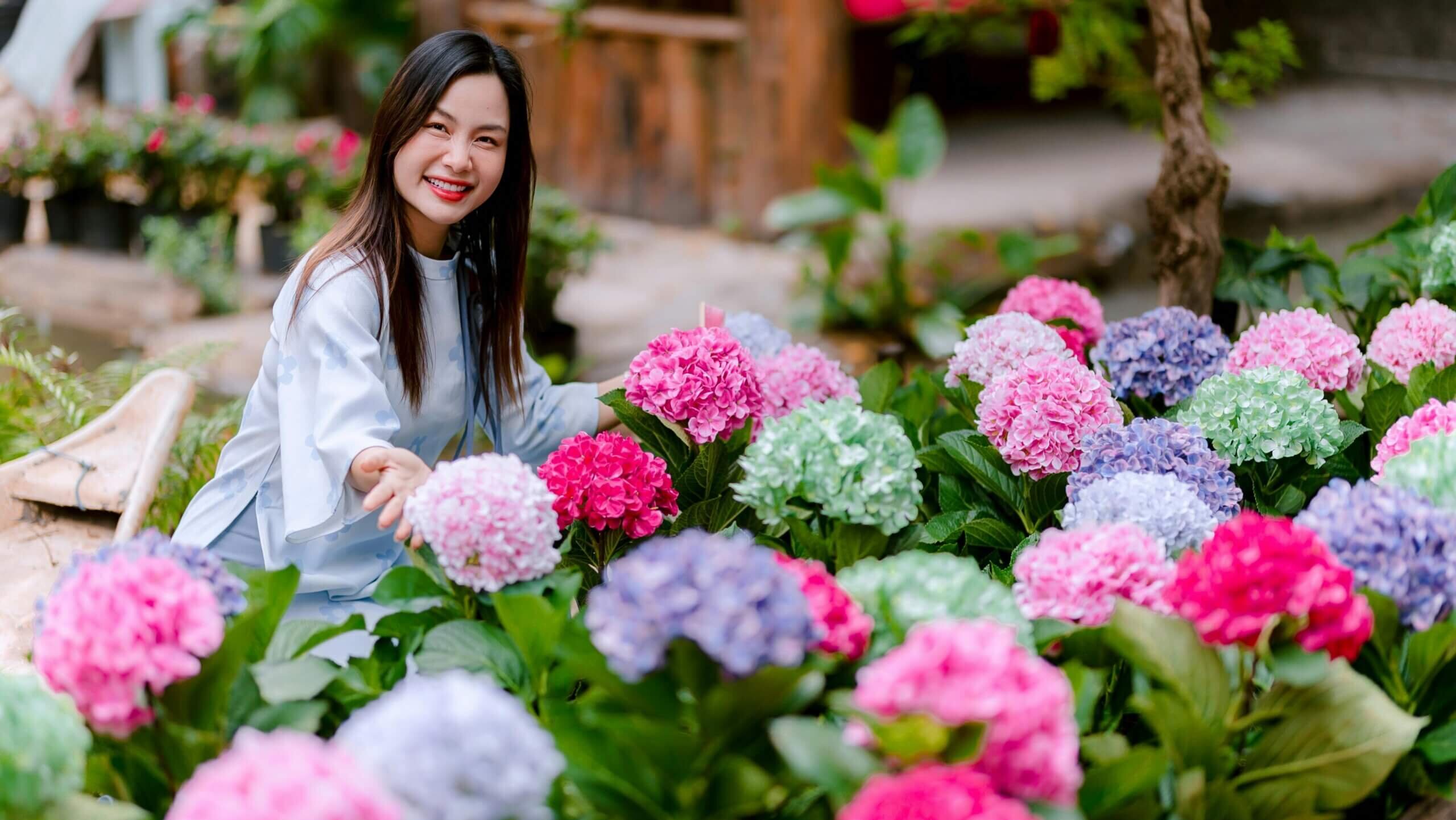 Smiling woman in light blue dress surrounded by colorful hydrangea flowers in a garden setting