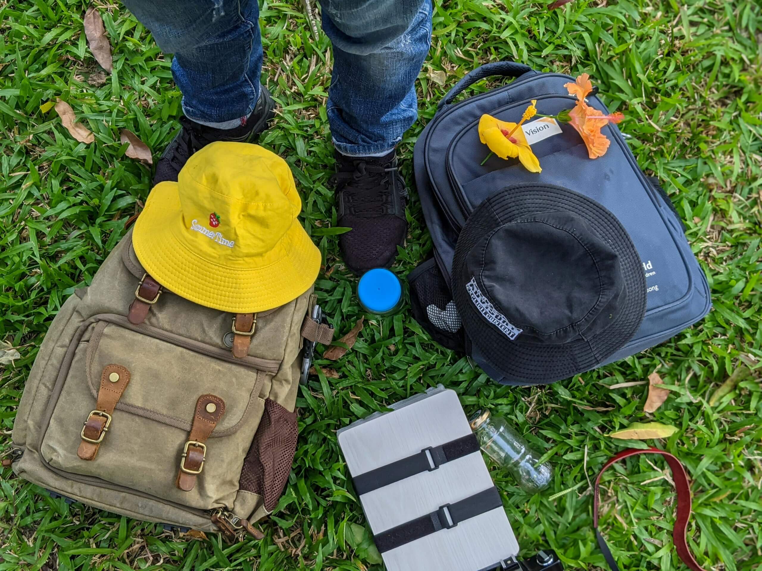 Person wearing jeans and black shoes standing on grass near two backpacks, yellow and black hats, flowers, and a water bottle.