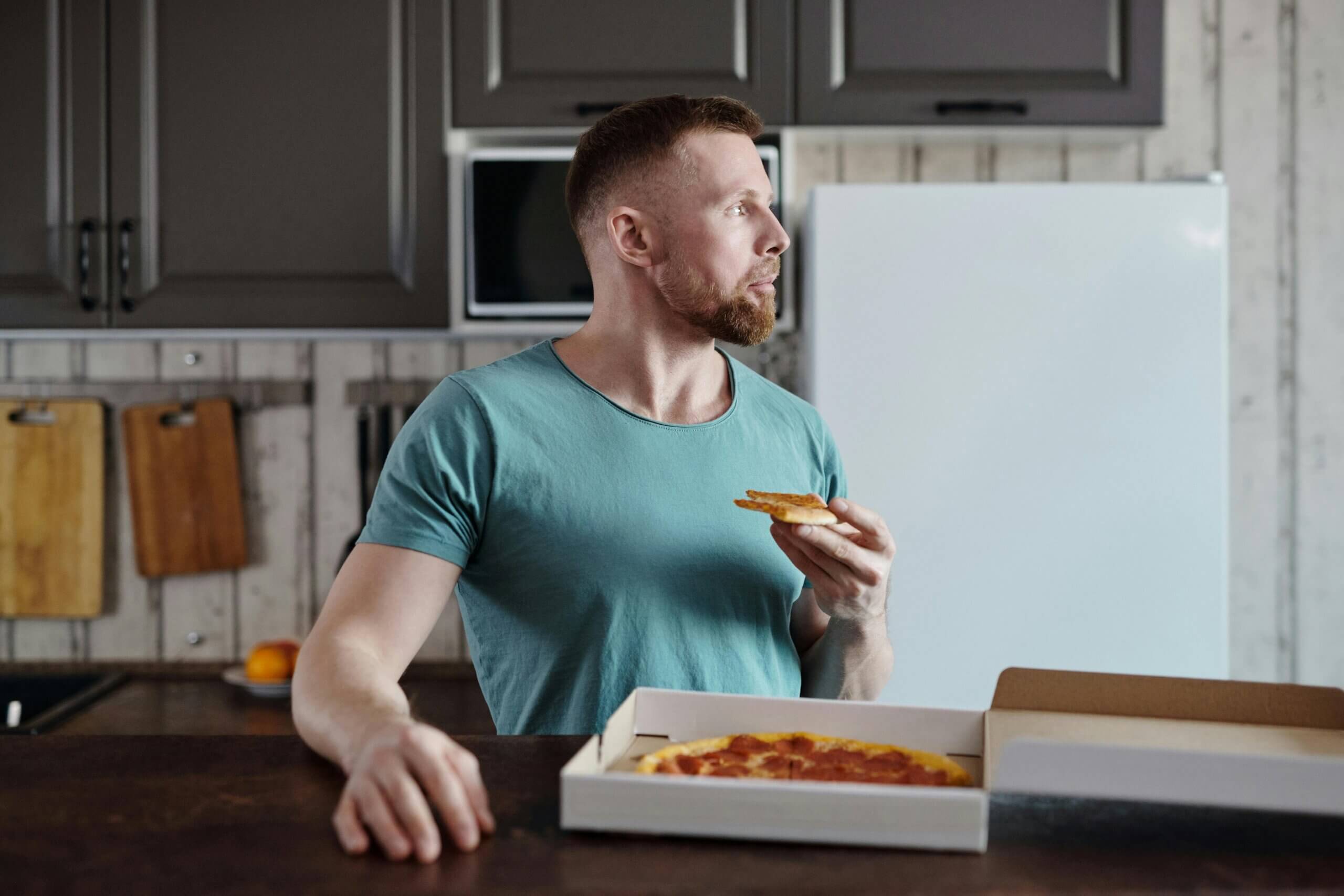 Man in teal shirt eating a slice of pepperoni pizza in a kitchen with wooden cabinets and cutting boards