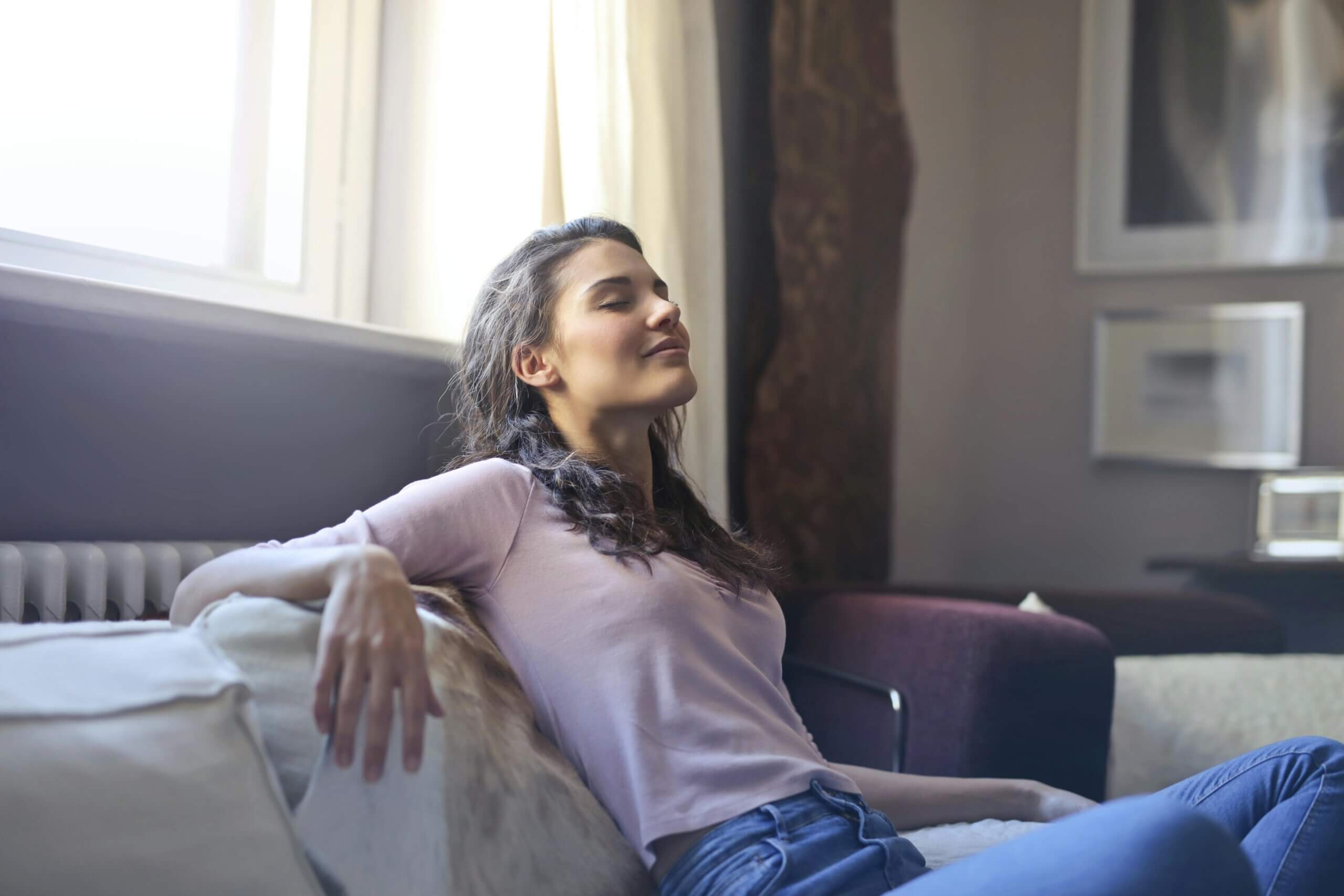 Young woman in a pink shirt and jeans relaxing on a couch with eyes closed near a window in a living room.