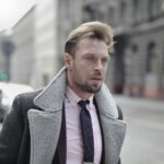 Man with styled hair wearing a gray shearling coat, pink shirt, and patterned tie walking on a city street