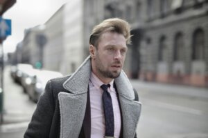 Man with styled hair wearing a gray shearling coat, pink shirt, and patterned tie walking on a city street