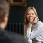 Smiling blonde woman in a white shirt resting her chin on her hand during a conversation with a man.