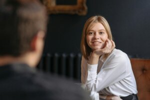 Smiling blonde woman in a white shirt resting her chin on her hand during a conversation with a man.