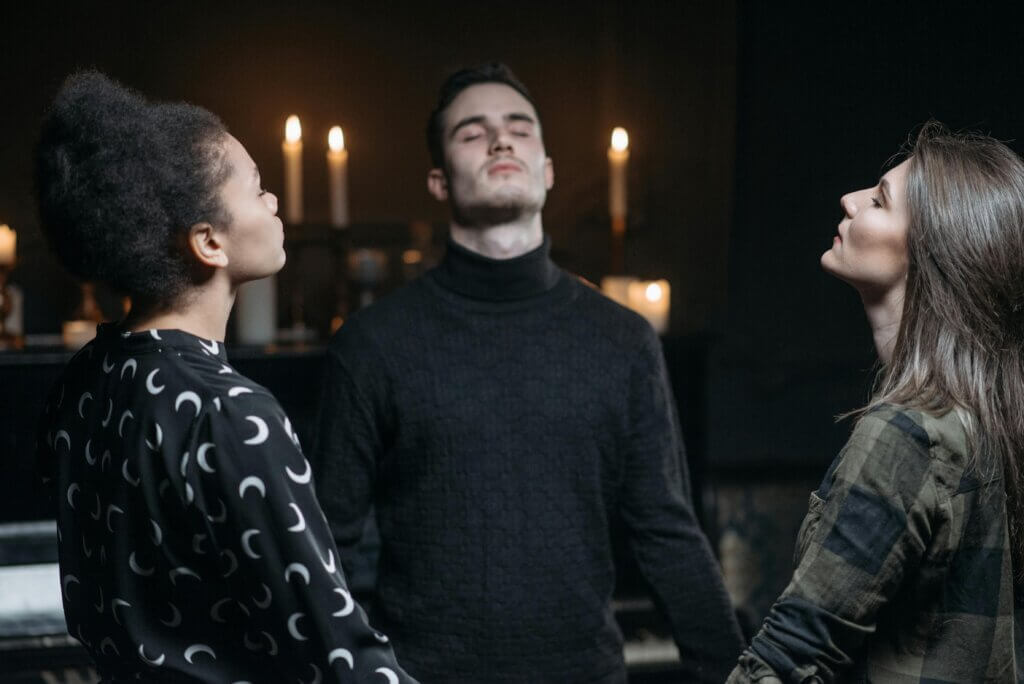 Three people standing in a circle with eyes closed, holding hands, with lit candles in the background.