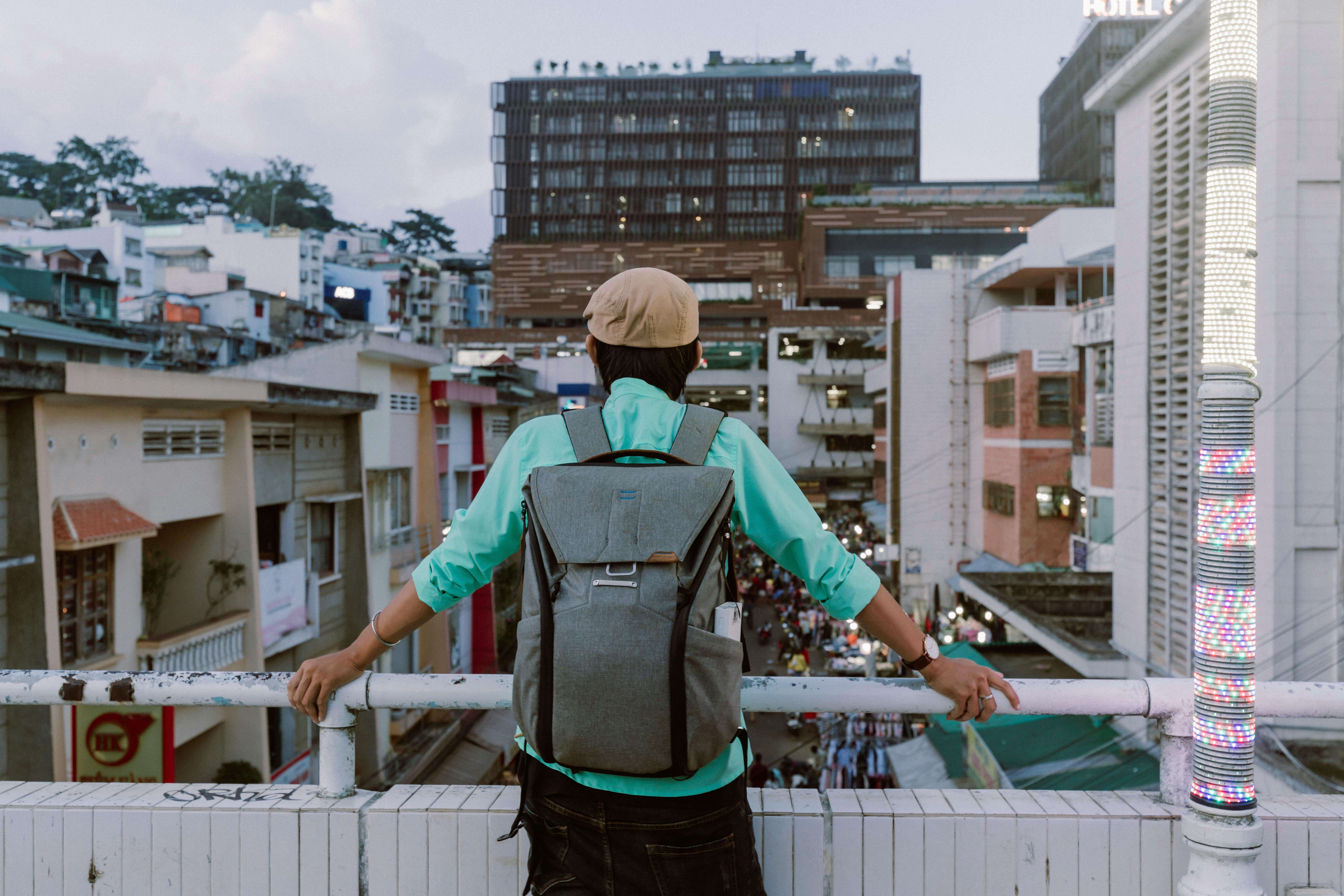 Person wearing a beige cap and teal shirt with a gray backpack overlooking a busy urban street from a balcony railing.