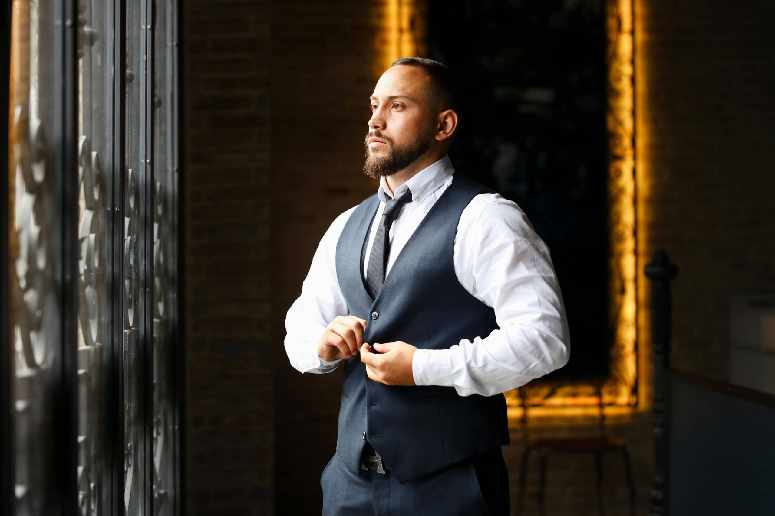 Man with beard buttoning navy vest over white shirt and black tie, standing by window in dimly lit room