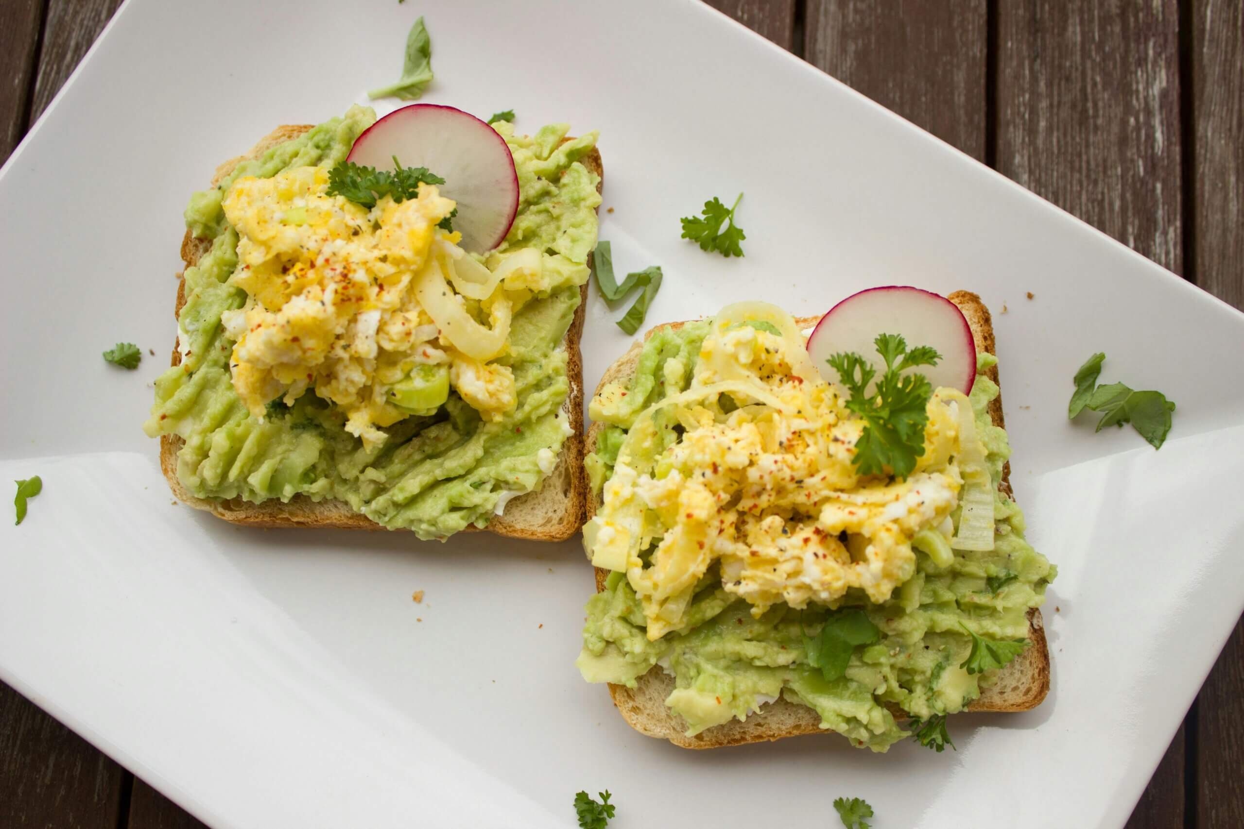Two slices of toast topped with mashed avocado, scrambled eggs, radish slices, and parsley on a white plate