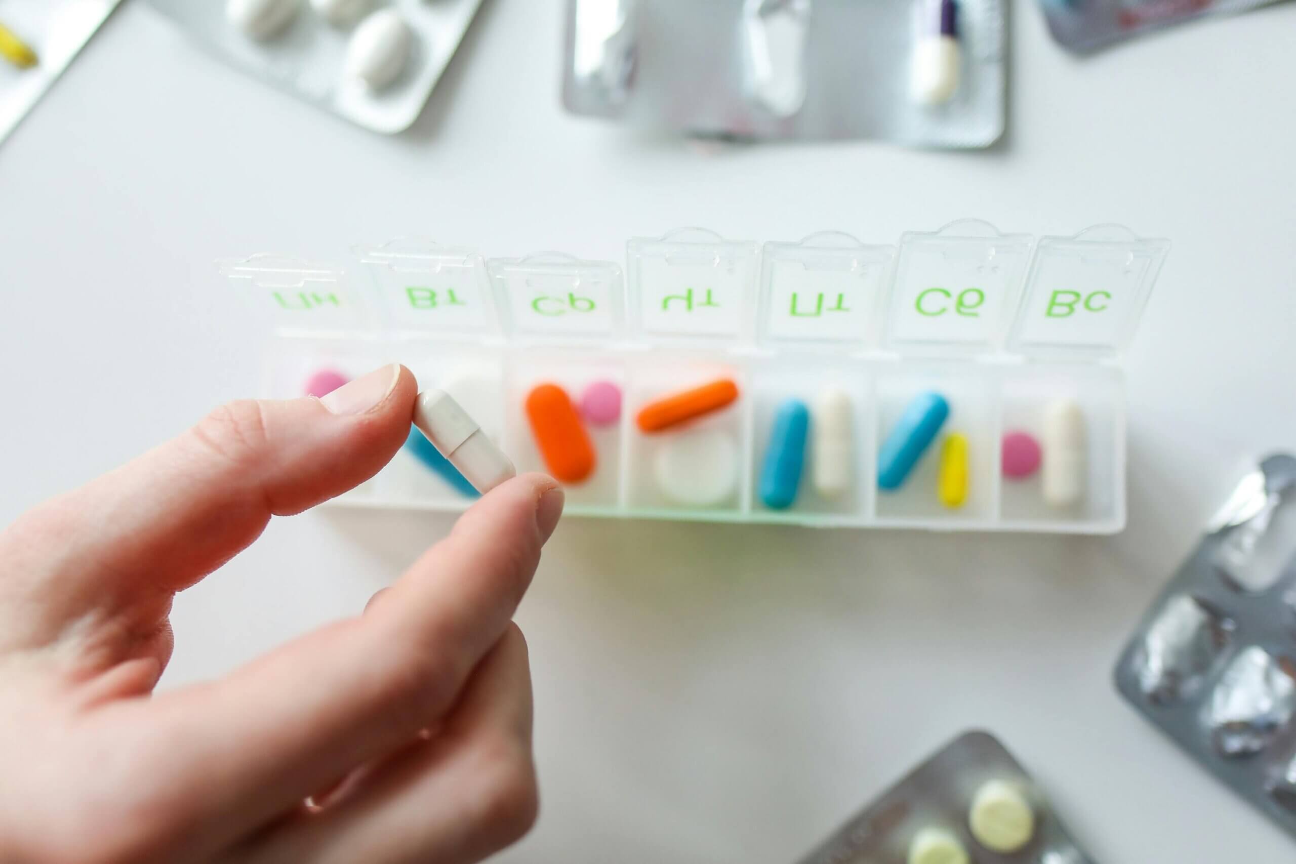 Hand holding a white capsule pill above a weekly pill organizer with various colored pills inside