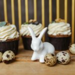 White ceramic rabbit figurine surrounded by speckled quail eggs and cupcakes with cream frosting on a wooden table