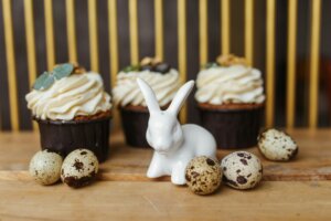 White ceramic rabbit figurine surrounded by speckled quail eggs and cupcakes with cream frosting on a wooden table