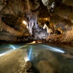 Underground cave with a flowing river illuminated by multiple underwater lights.