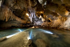 Underground cave with a flowing river illuminated by multiple underwater lights.