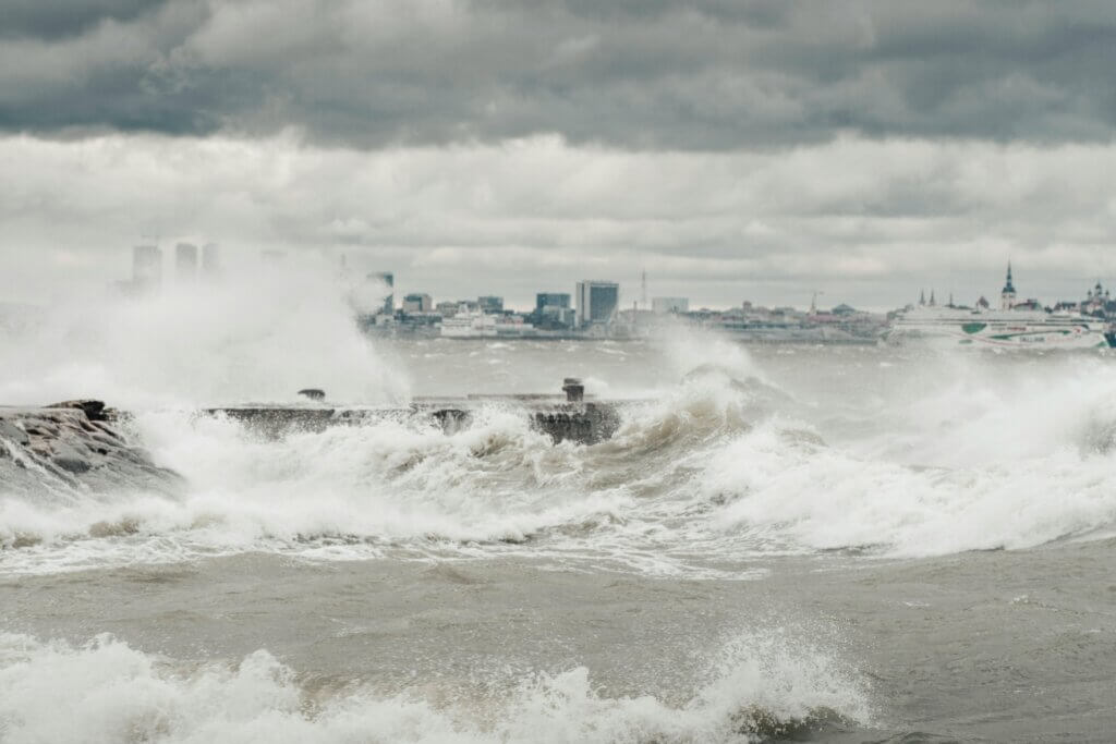 Rough sea waves crashing against a pier with a city skyline and cloudy sky in the background.