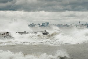 Rough sea waves crashing against a pier with a city skyline and cloudy sky in the background.