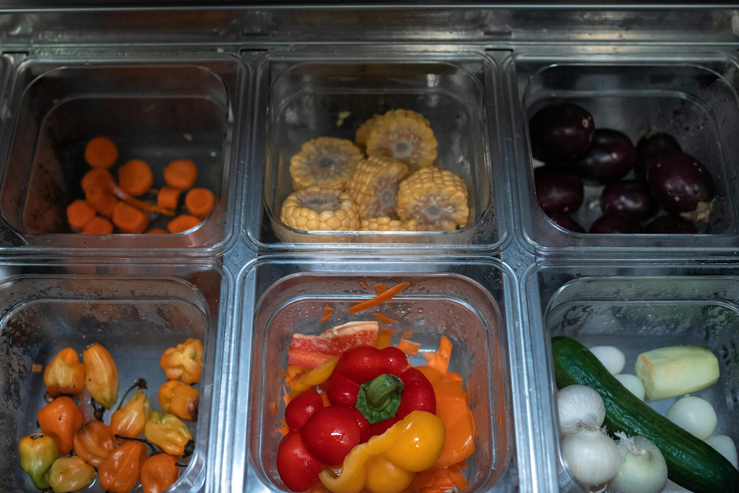 Containers with sliced carrots, corn, plums, habanero peppers, bell peppers, garlic, cucumber, and onions in a salad bar.
