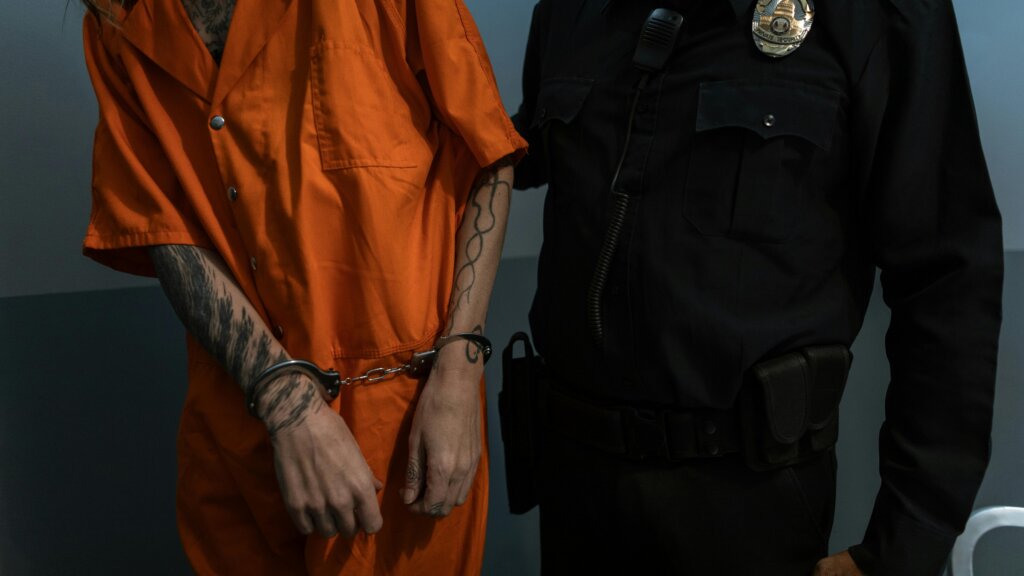 Person in orange prison uniform with handcuffs escorted by security officer in black uniform