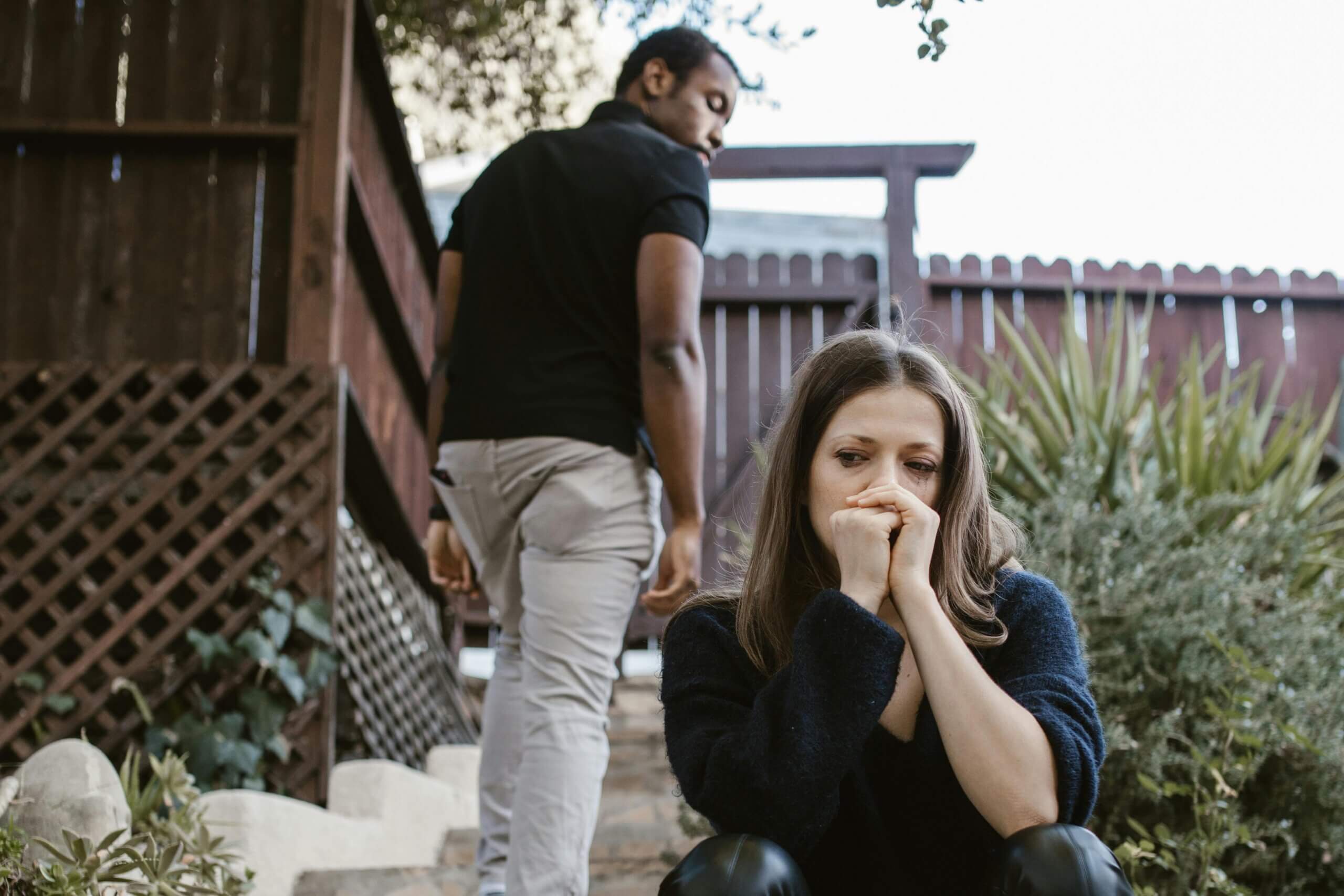 Woman sitting with hands near mouth looking upset while man walks away in a backyard setting