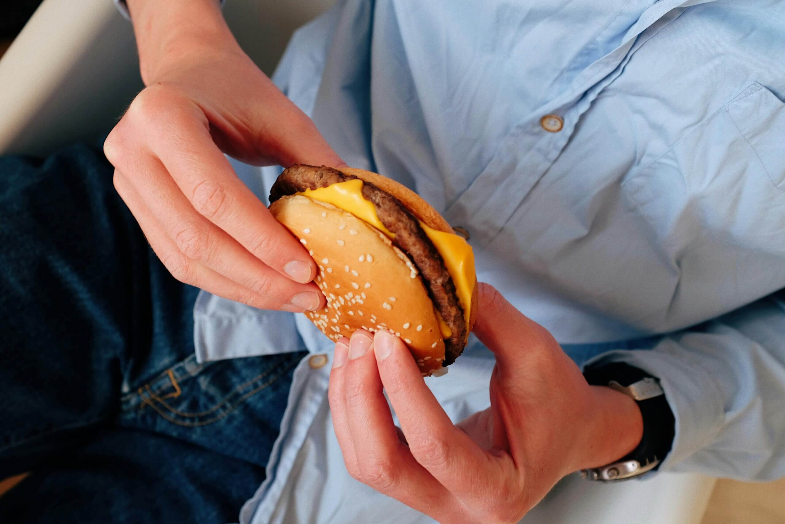 Person in blue shirt holding a cheeseburger with sesame seed bun and melted cheese