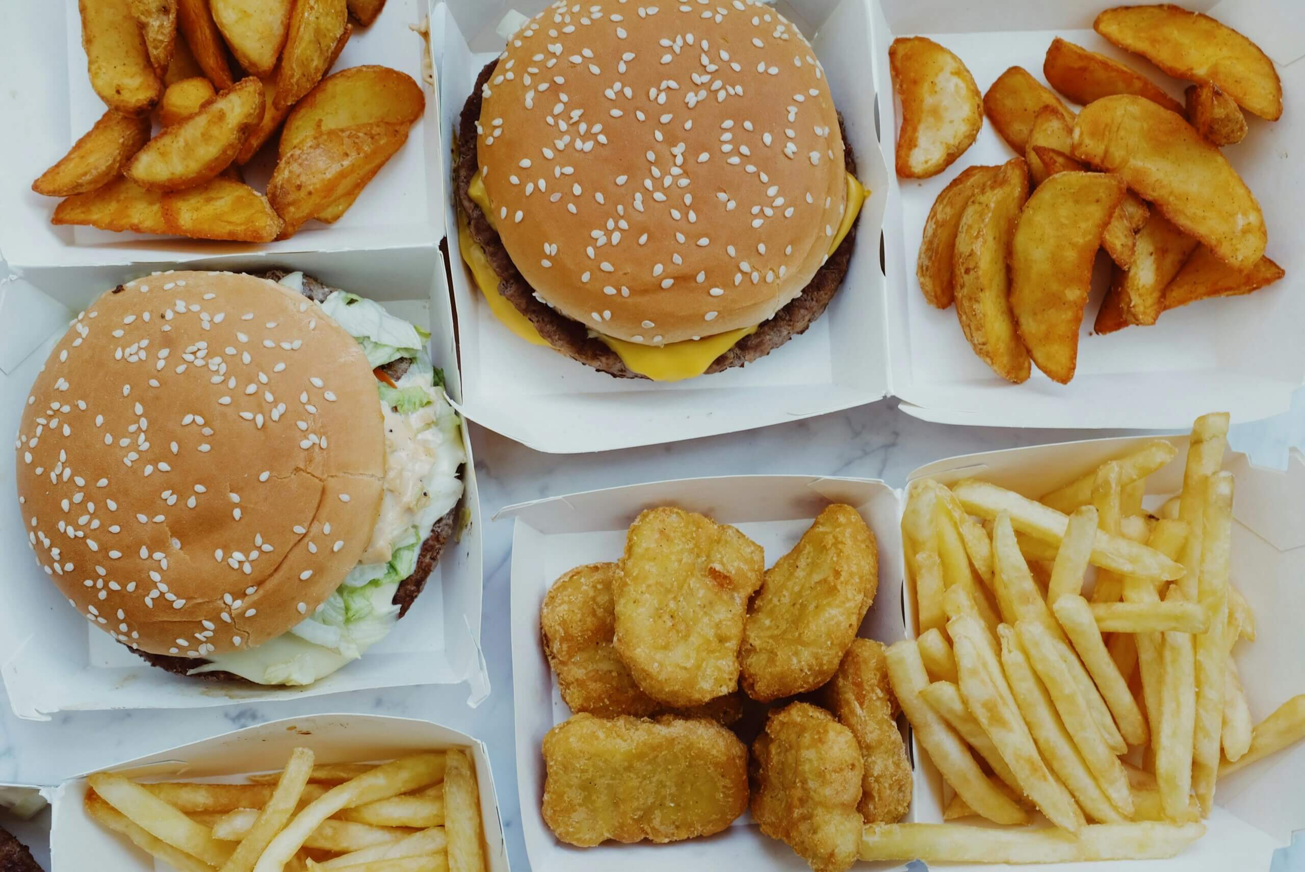 Two cheeseburgers, potato wedges, chicken nuggets, and French fries in white fast food containers on a marble surface
