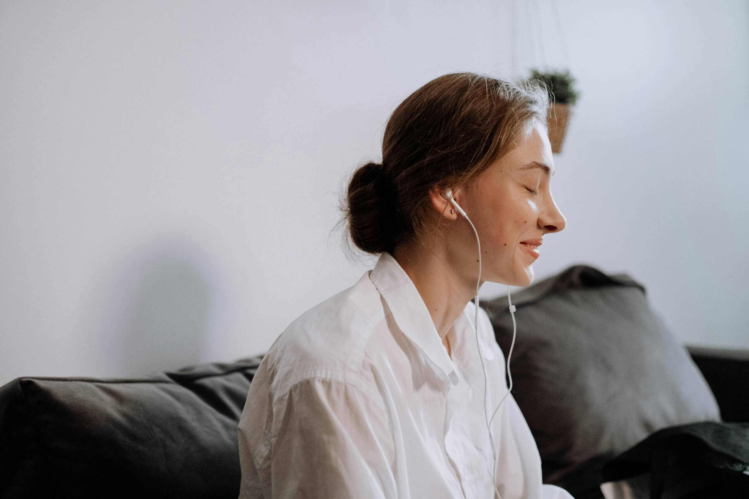 Young woman with earbuds smiling with eyes closed while sitting on a couch in a white shirt
