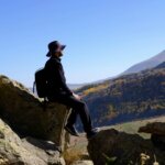 Man in black hiking outfit and hat sitting on rocks overlooking a mountainous landscape with autumn foliage.